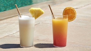 Duas bebidas com canudos e frutas em uma mesa à beira da piscina, com piscina e um edifício ao fundo.
