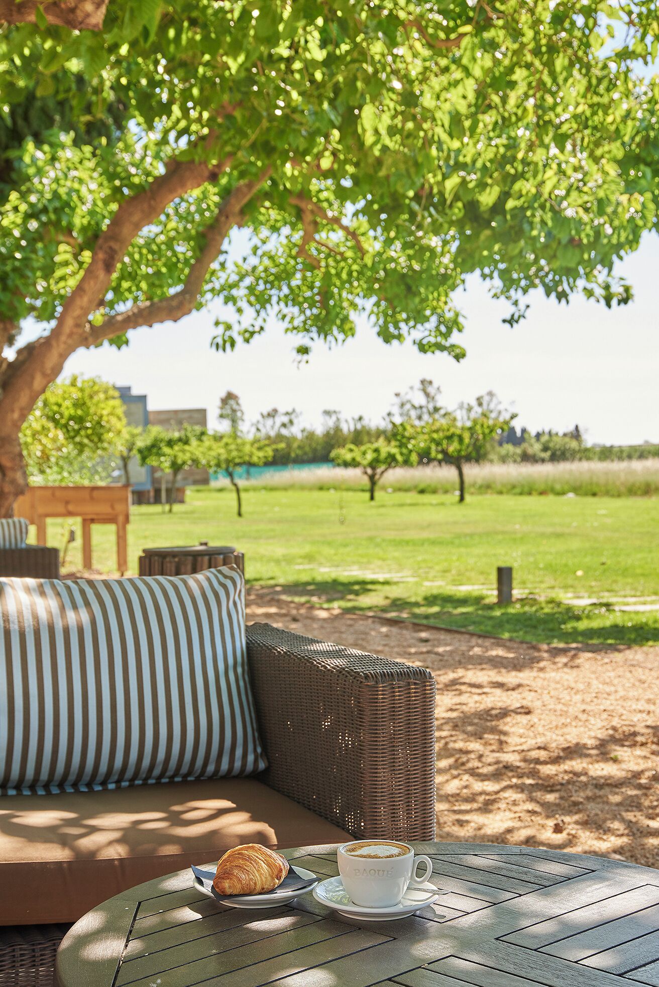 Un espacio al aire libre con un sofá de mimbre, almohadones a rayas y una mesa con un croissant y una taza de café.