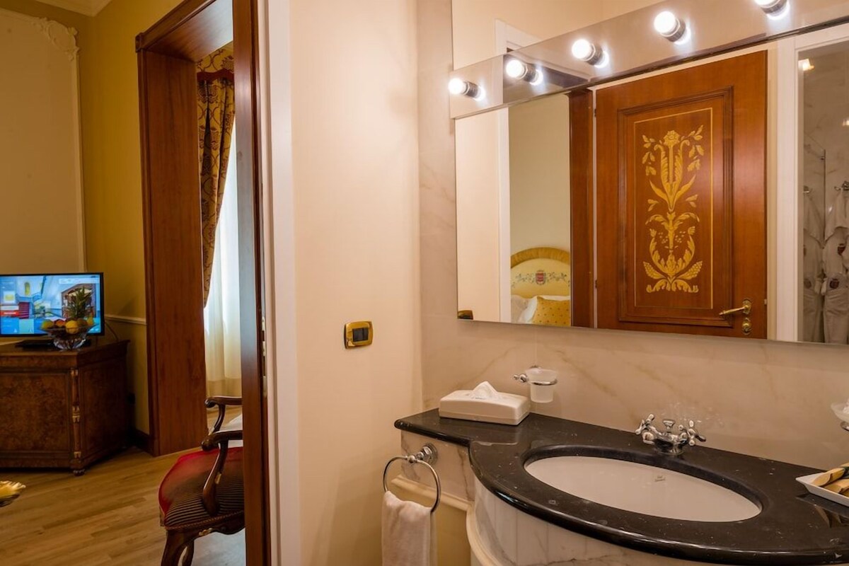 Un baño con un lavamanos negro, un espejo con luces, una puerta de madera con un emblema dorado y un televisor sobre un mueble de madera.