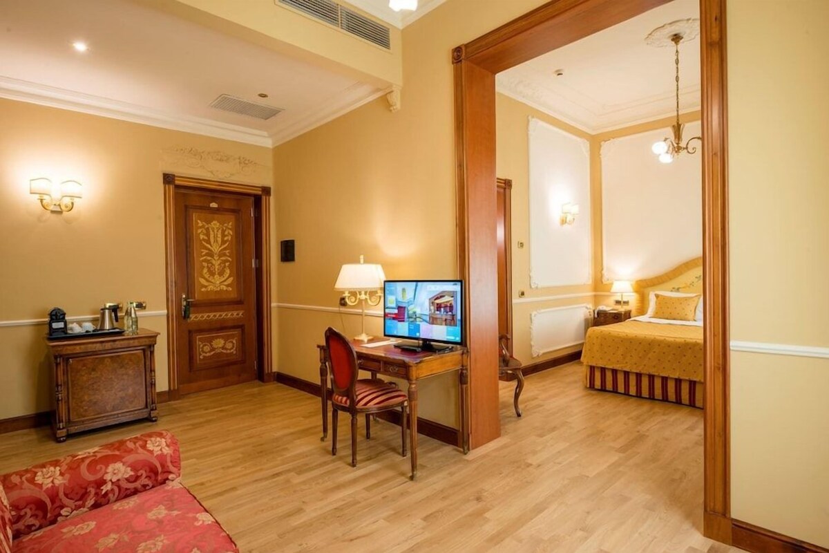 Hotelkamer met een houten bureau, een computer, een bed met een gele sprei, een rode gestoffeerde bank en een houten deur met decoratief paneel.
