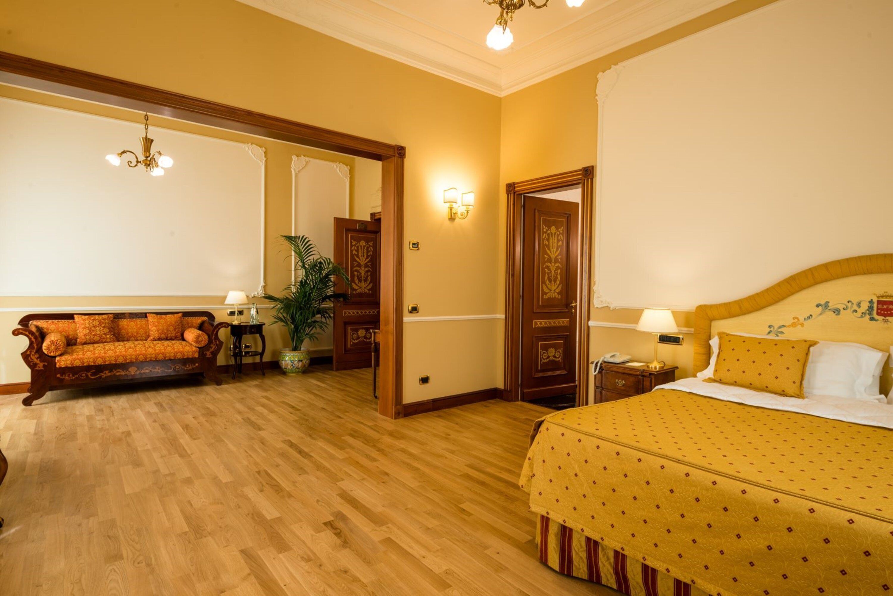 Een hotelkamer met een houten vloer, een bank, een bed met een gele plaid, een nachtkastje, een lamp en een wandlamp.