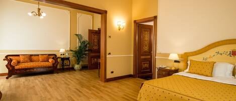 Habitación de hotel con piso de madera, un sofá, una cama con colcha amarilla, una mesita de noche, una lámpara y un aplique de pared.