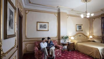 Ein Zimmer mit rotem Teppich, einem Bett mit goldenem Kopfteil, einer Sitzecke mit Sofa und gerahmten Bildern an den Wänden.