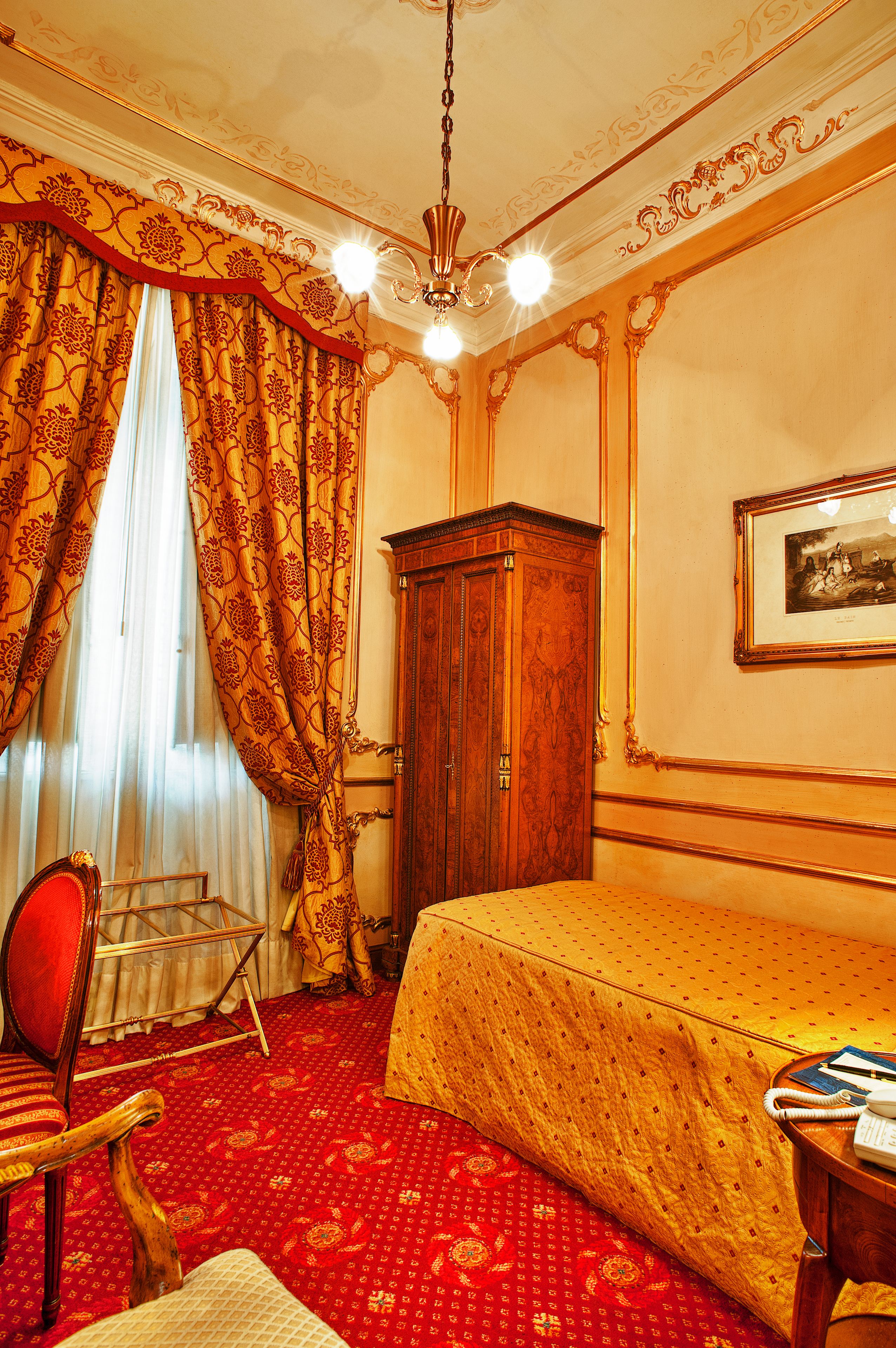 Una habitación de hotel con alfombra roja, un armario de madera, una cama con colcha amarilla, un sillón rojo y una lámpara de araña.