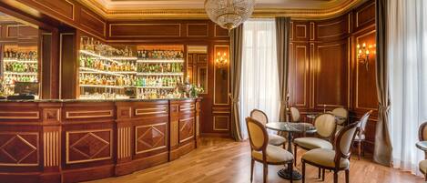 A bar area with a wooden bar and shelves of bottles, a chandelier, and a dining area with chairs and a table.
