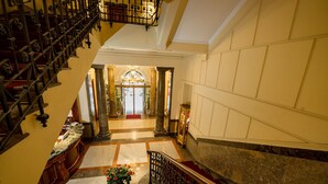 Una imponente escalera con barandillas ornamentales que conduce a un amplio vestíbulo con una araña y una alfombra roja.