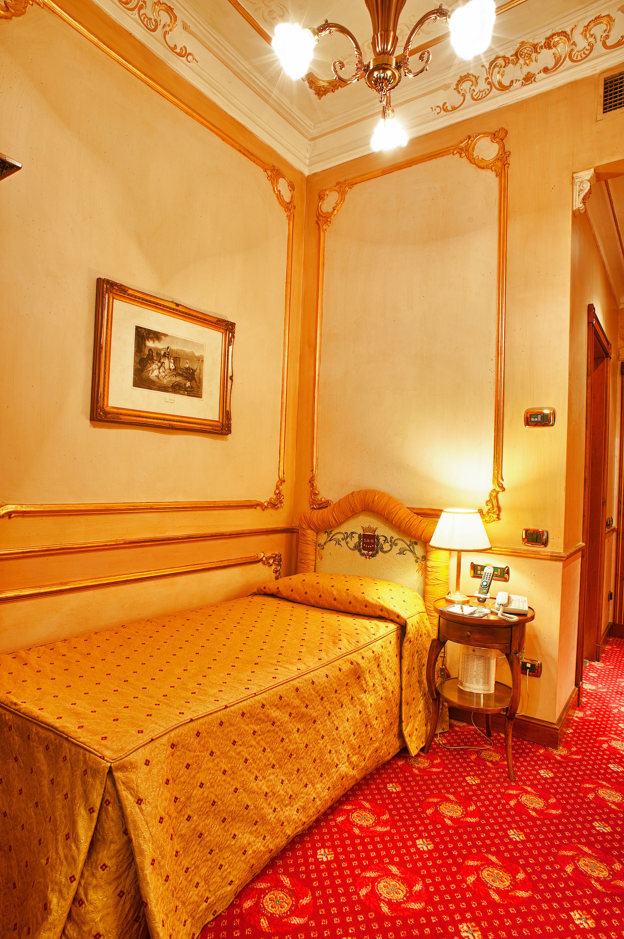 Une chambre d’hôtel avec un lit, une table de chevet, une lampe, un tableau encadré au mur et un tapis rouge à motifs.