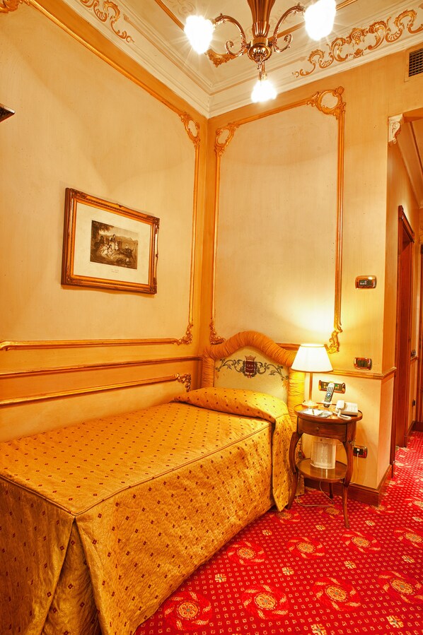 Habitación de hotel con cama, mesita de noche, lámpara, un cuadro en la pared y alfombra roja estampada.