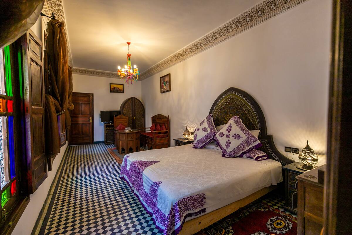 A traditional room with a checkered floor, a bed with a headboard, a chandelier, and a wooden cabinet.