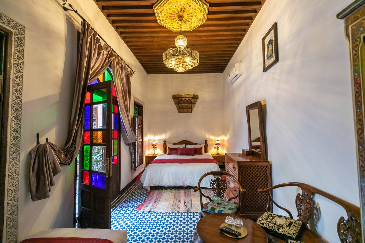 A room with a bed, a wooden chair, a table with a tray, and a chandelier.