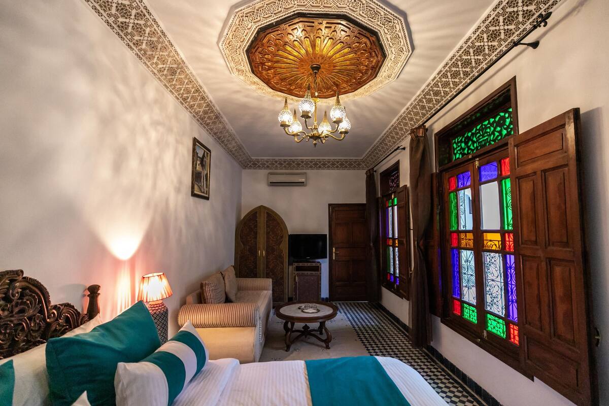 A room with a bed, a sofa, a table, a chandelier, and a stained glass window.