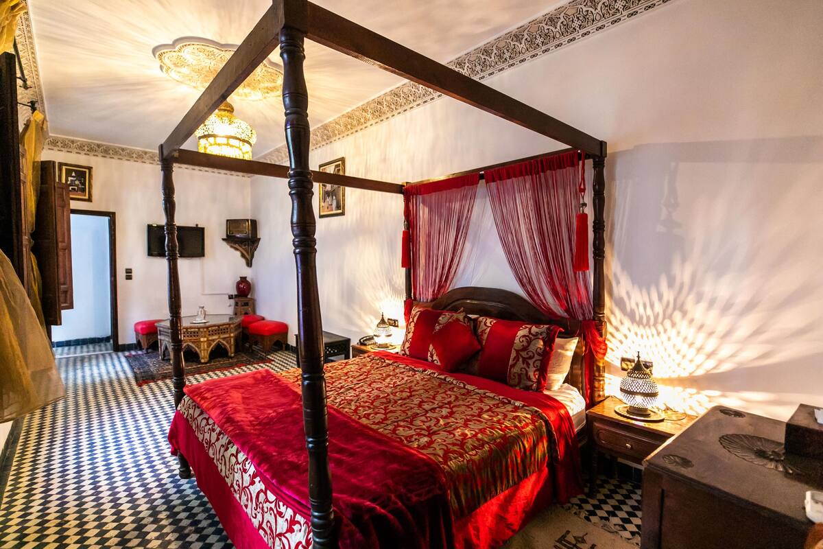 A four-poster bed with red and gold bedding, a checkered floor, and a chandelier.