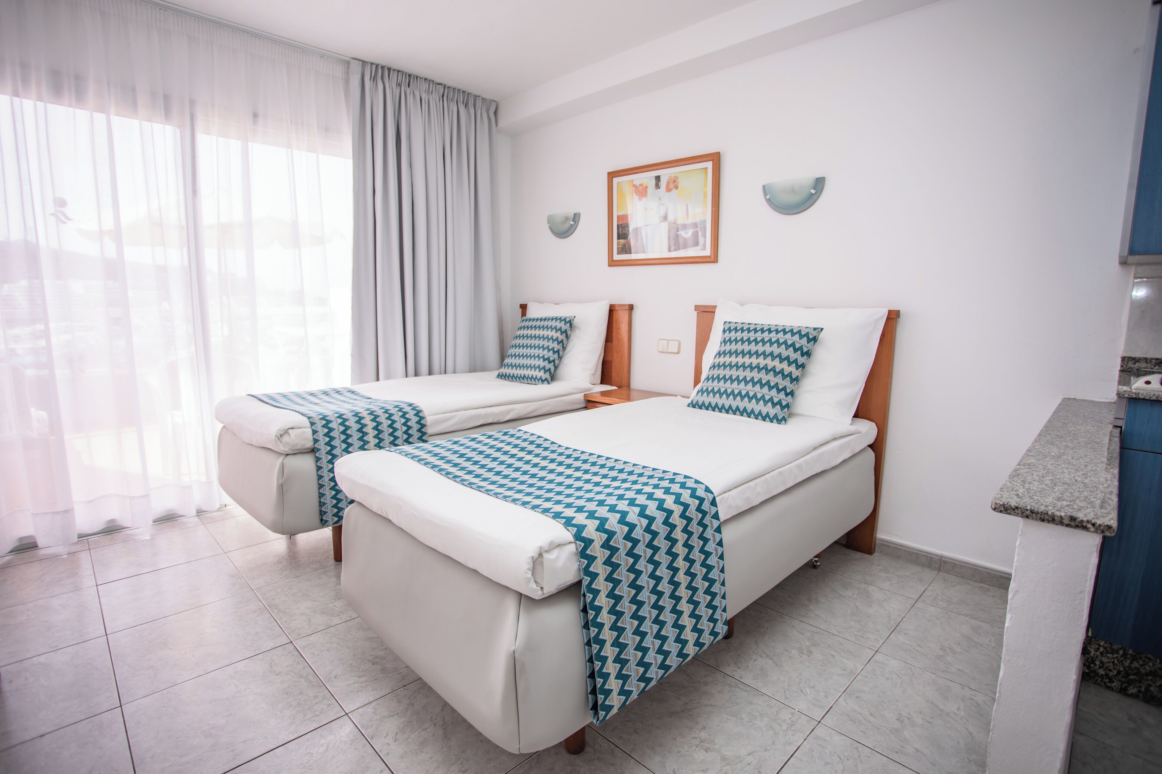 A hotel room with two beds, a small kitchenette, and a large window with curtains.