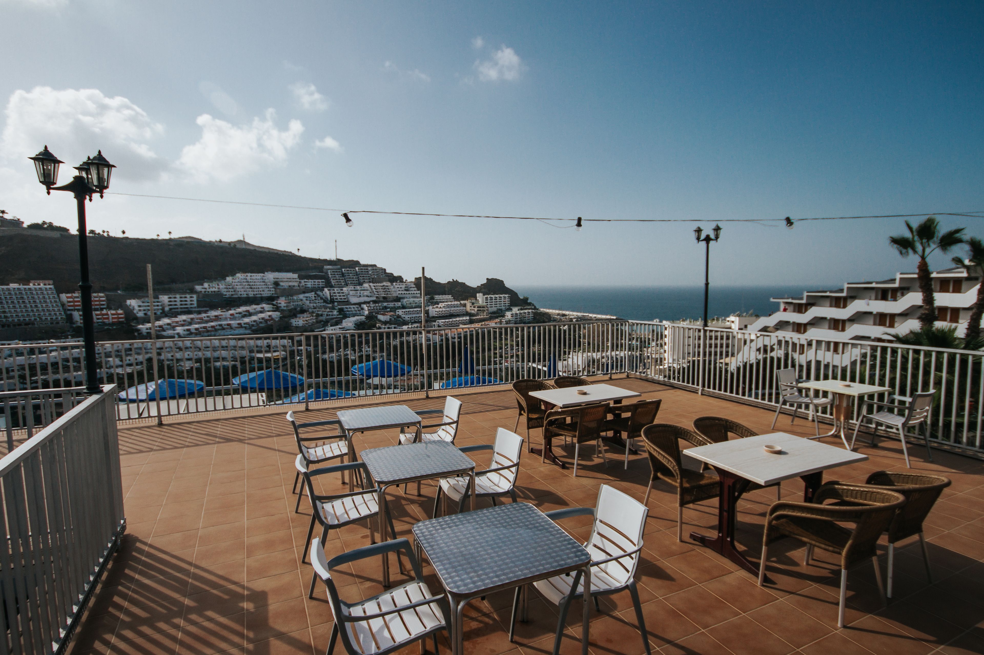 A terrace with tables and chairs, overlooking a coastal town and the sea.