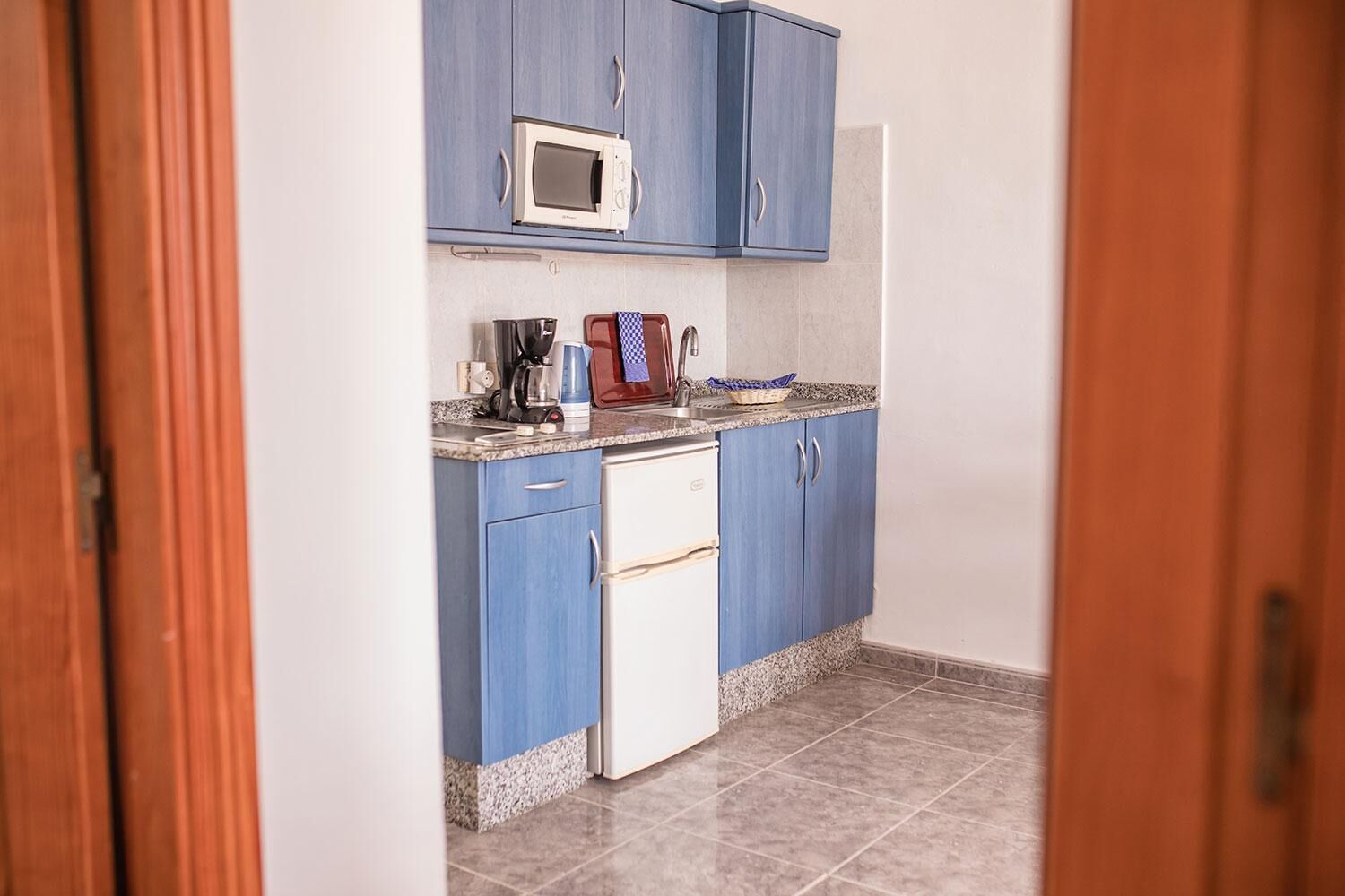 A compact kitchen with blue cabinets, a microwave, a coffee maker, and a small refrigerator.