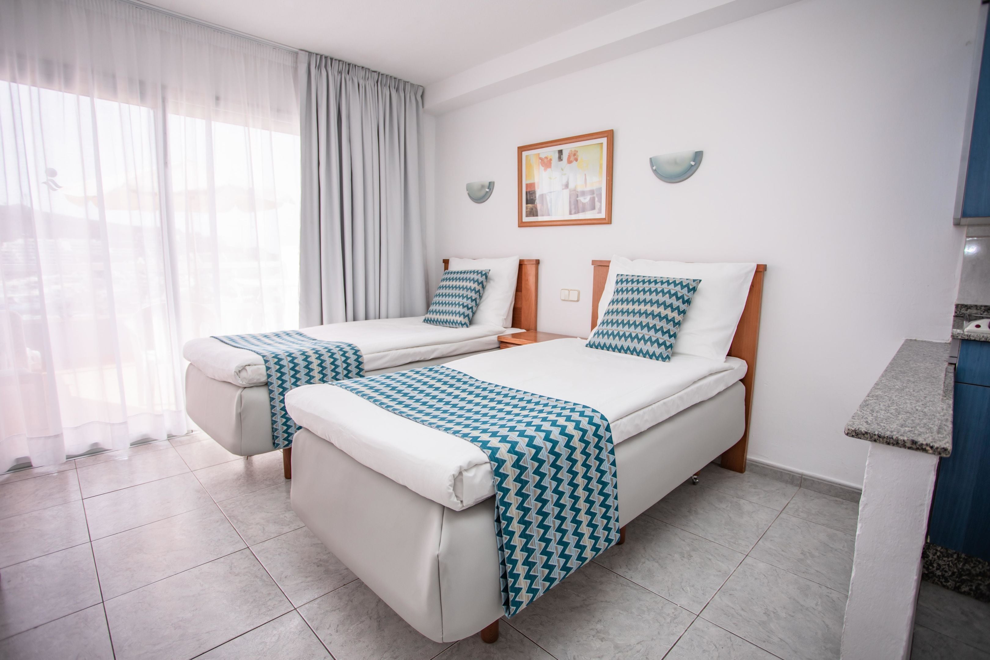 A hotel room with two beds, a small kitchenette, and a large window with sheer curtains.