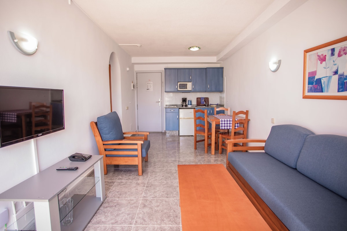 Un espacio habitable compacto con televisor, sofá, sillas, una pequeña cocina y una mesa de comedor.
