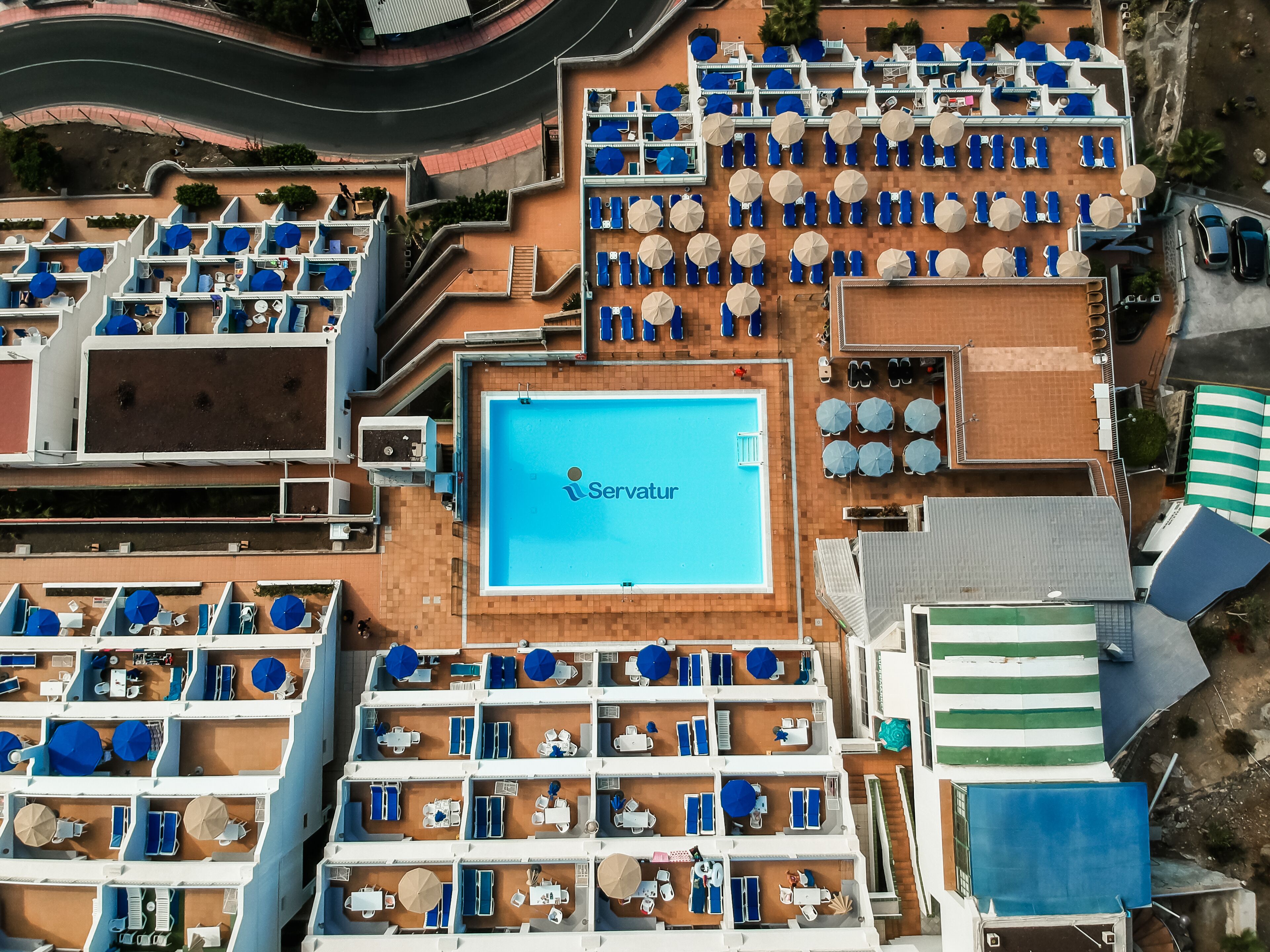 Vista aérea de un hotel con varios pisos, piscina y numerosos camastros y sombrillas.
