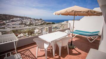 Un balcon avec une table et des chaises blanches, un parasol beige et une chaise bleue avec un coussin.
