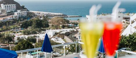 A balcony with lounge chairs and umbrellas overlooking a coastal town with buildings and a view of the sea.