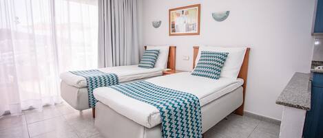 Ein Hotelzimmer mit zwei Betten, einer kleinen Küchenzeile und einem großen Fenster mit durchsichtigen Vorhängen.