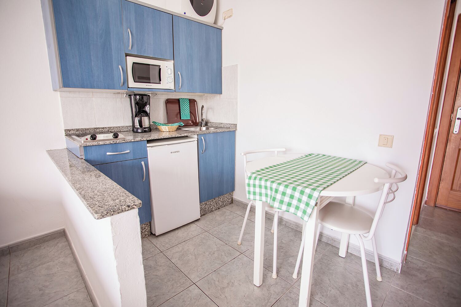 Une cuisine compacte avec des placards bleus, un four à micro-ondes, une machine à café et une petite table recouverte d’une nappe à carreaux.