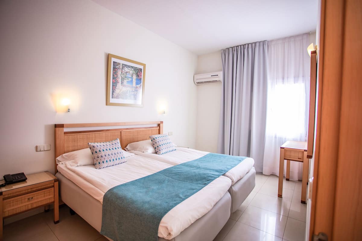 A hotel room with a large bed, bedside tables, an air conditioning unit, a window with curtains, and a small table.