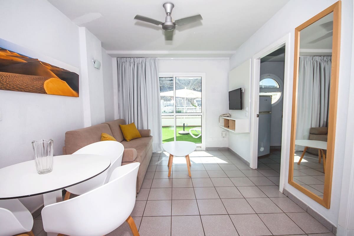 A modern living room with a sofa, a dining table, and a TV. There is a balcony with a view of greenery and buildings.