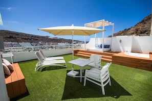 Una terraza en la azotea con vistas a un pueblo costero, que incluye un área de estar con mesa y sillas, un toldo blanco y un pequeño rincón de asiento.