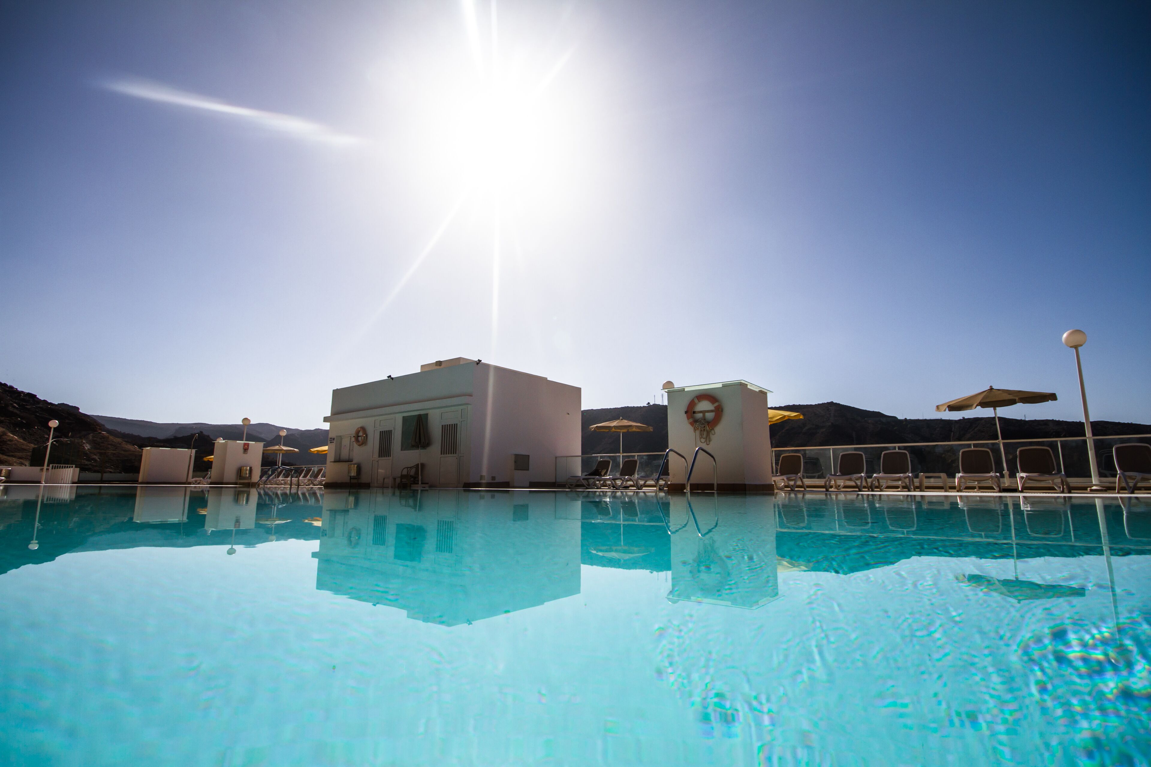 A clear blue pool with sun reflecting on the water, surrounded by lounge chairs and umbrellas, with a mountainous backdrop.