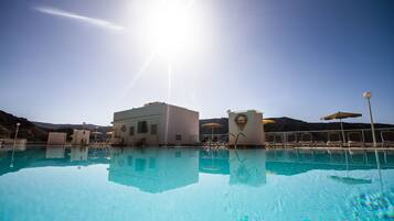 A clear blue pool with sun reflecting on the water, surrounded by lounge chairs and umbrellas, with a mountainous backdrop.