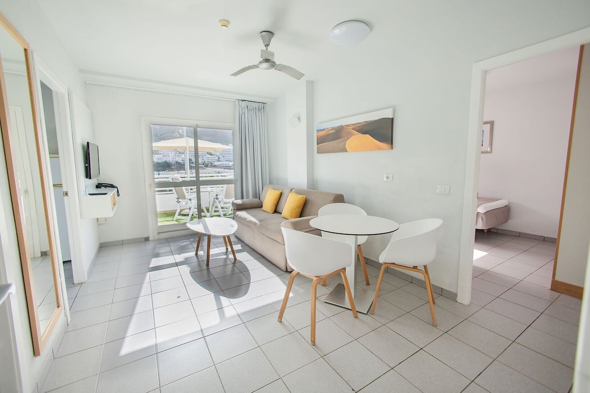 A modern living room with a sofa, two white chairs, a small table, a ceiling fan, and a large window with a view of buildings.