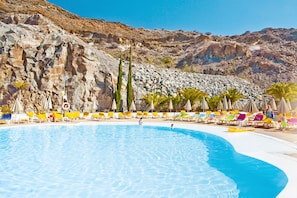Una piscina con agua cristalina azul, rodeada de sillas de descanso y sombrillas coloridas, y colinas rocosas al fondo.