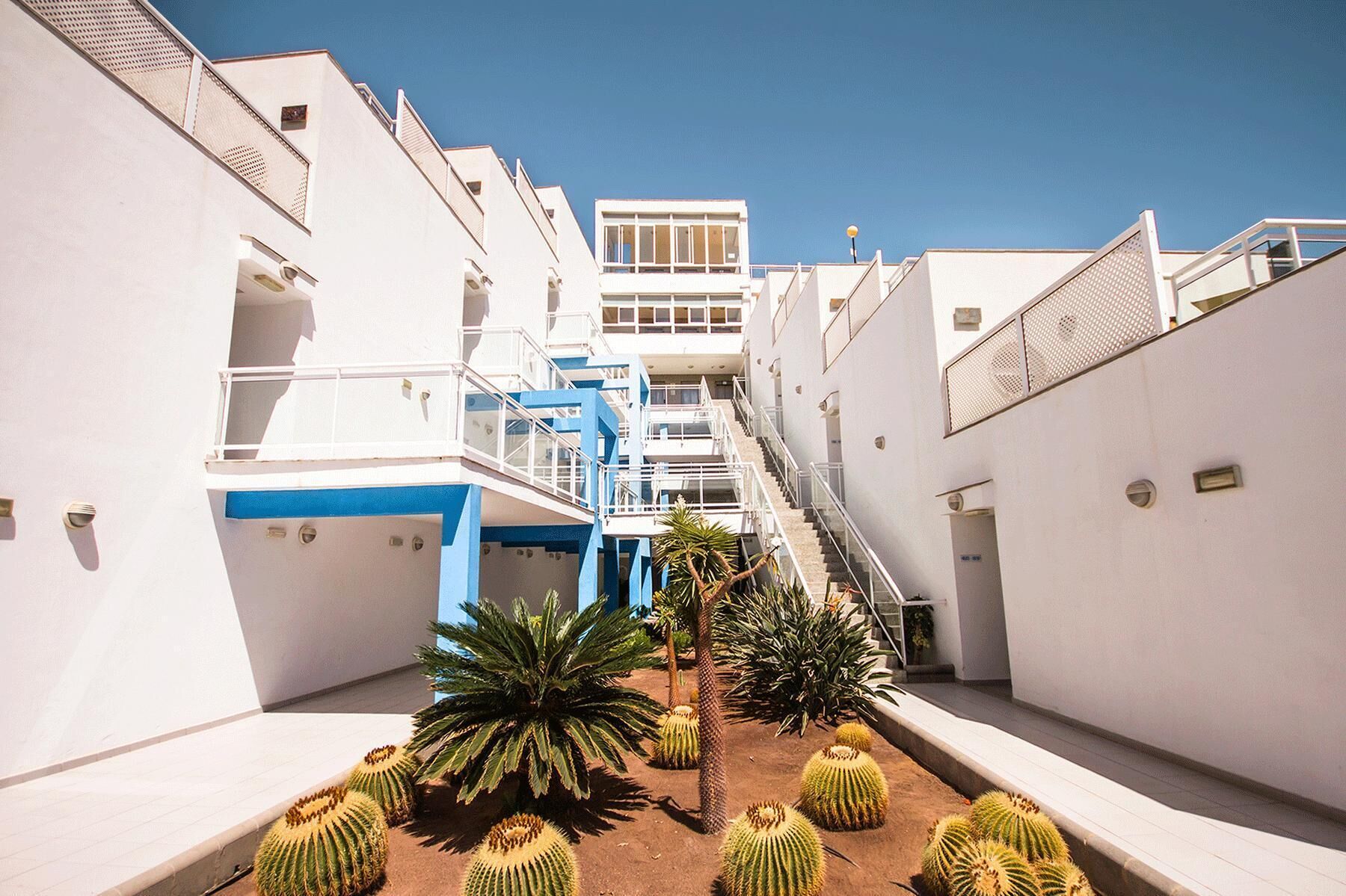 Un hôtel moderne doté d’une cour intérieure où l’on trouve des cactus et des palmiers, entouré de bâtiments blancs rehaussés d’accents bleus et agrémenté de nombreux balcons.