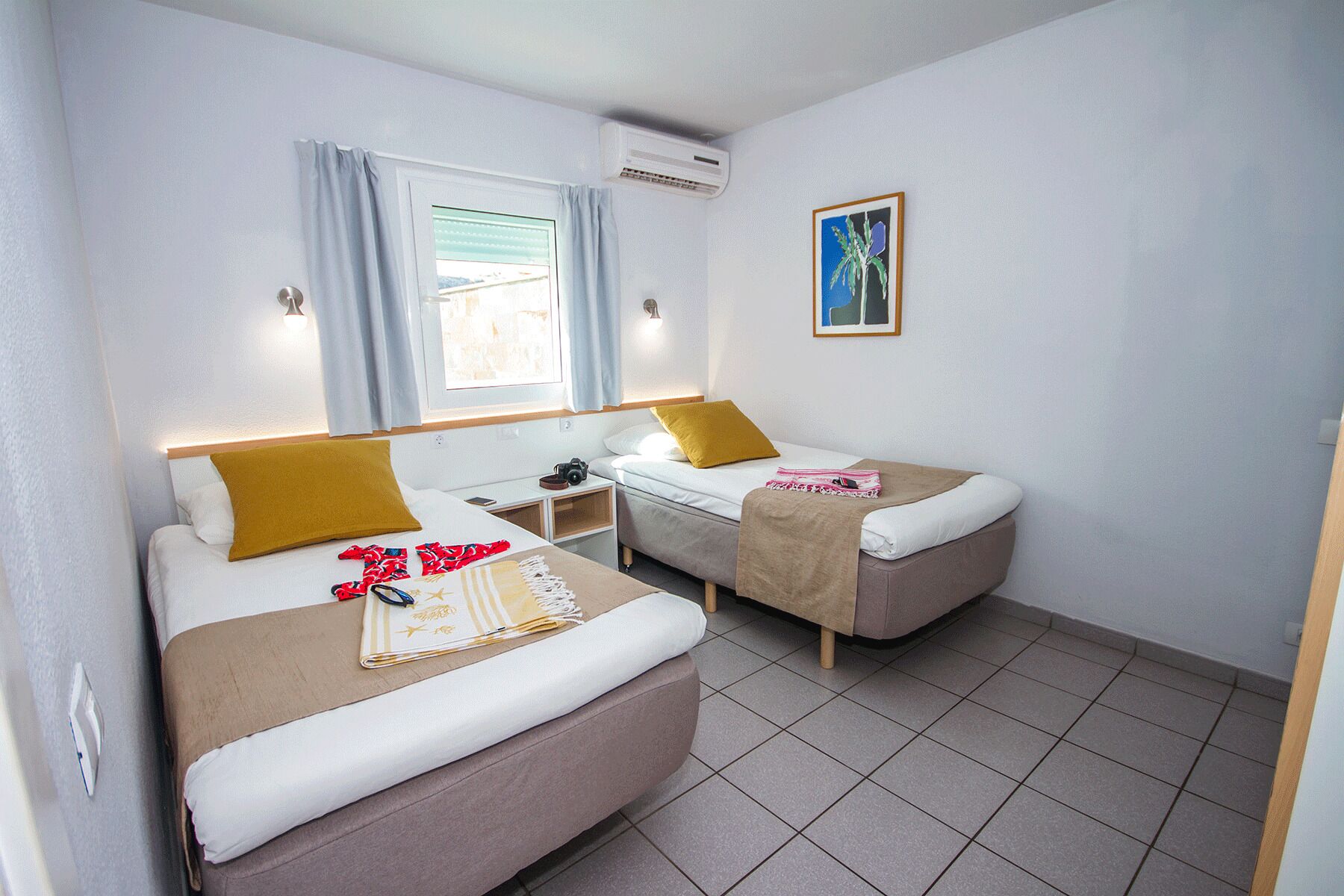 Habitación de hotel con dos camas, una ventana con cortinas, un equipo de aire acondicionado, una mesita de noche con una cámara y un cuadro en la pared.