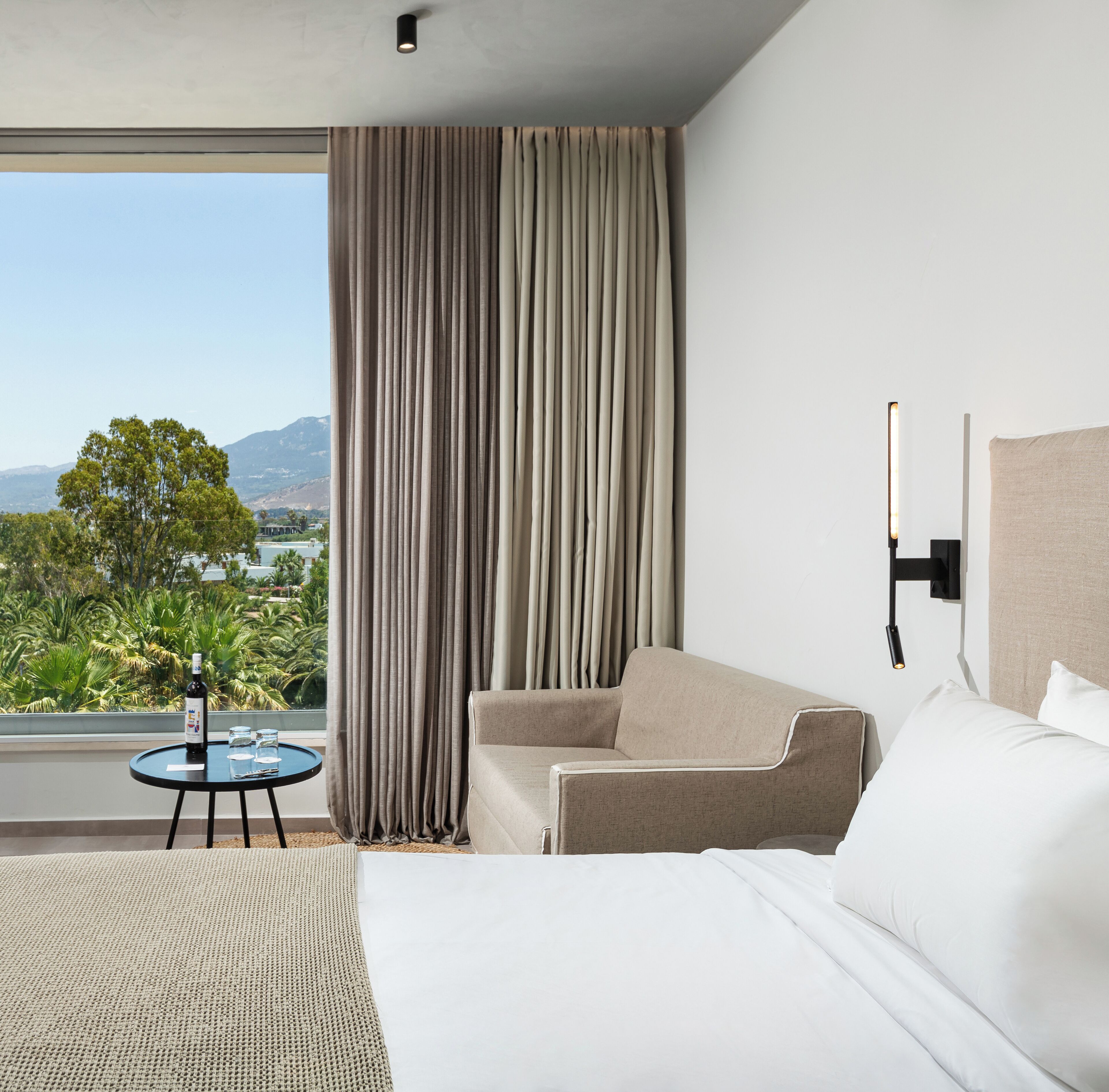 Een hotelkamer met een bed, een bank, een klein tafeltje met een fles en glazen, uitzicht op het omliggende landschap en een wandlamp.