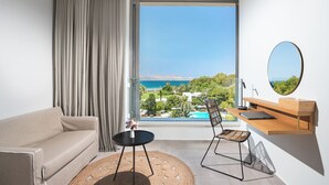 A modern room with a sofa, a small round table, a chair, a desk with a mirror, and a large window offering a view of the sea and greenery. - Caravia Beach Hotel & Bungalows - All Inclusive (Kos)