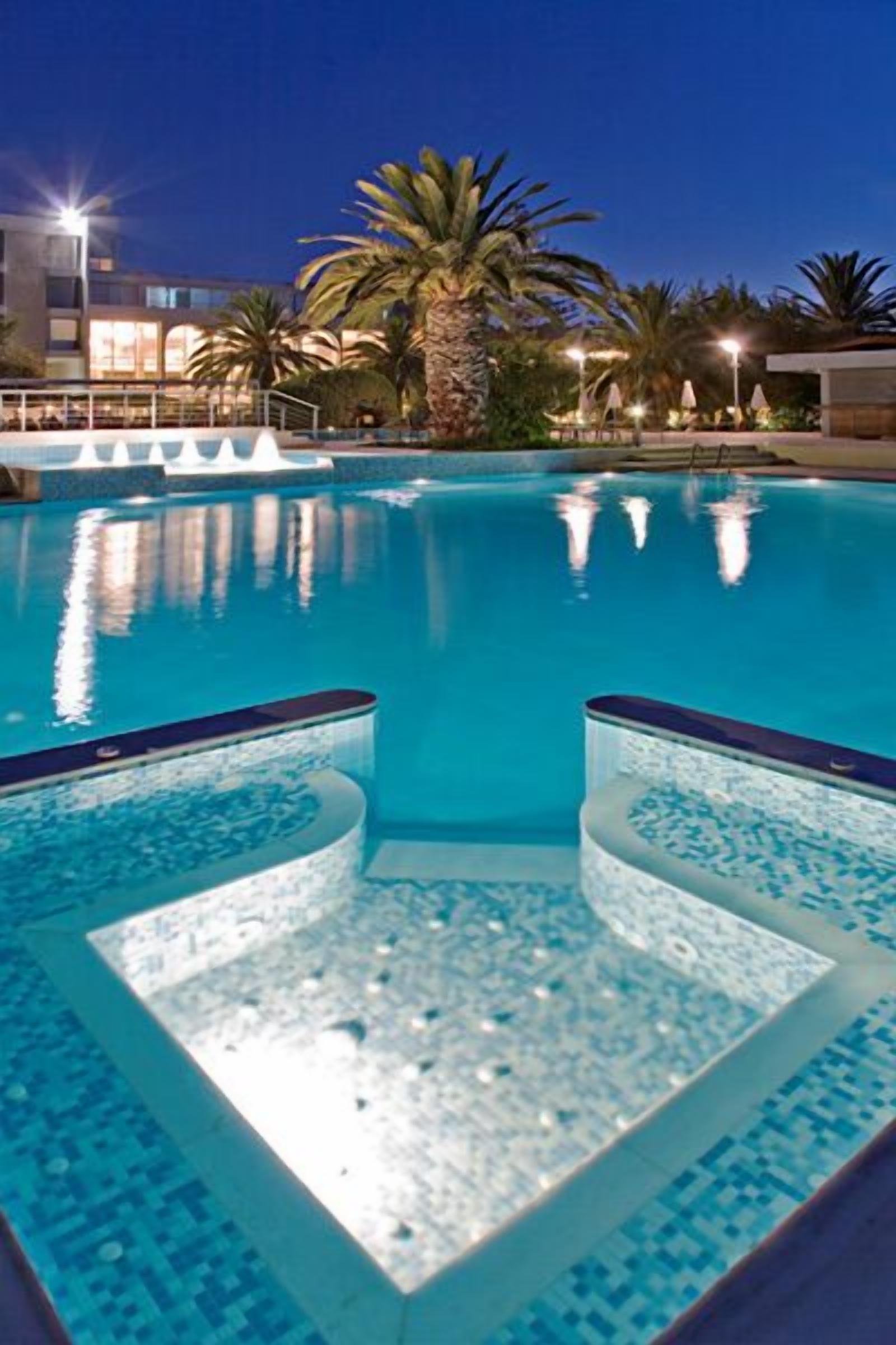Una piscina con agua azul cristalina, rodeada de palmeras y un edificio al fondo.