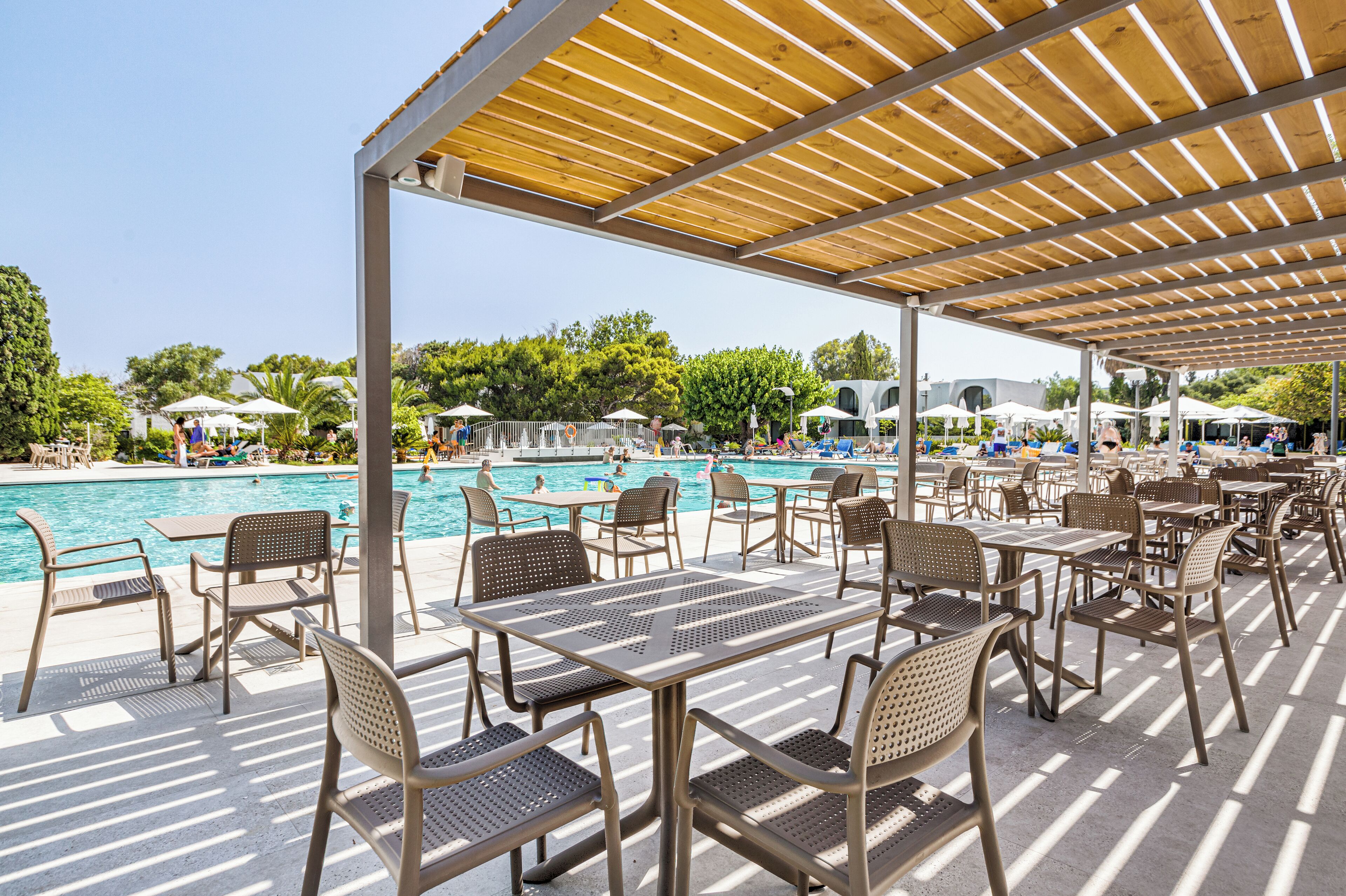 A poolside area with tables and chairs under a wooden pergola, overlooking a swimming pool.