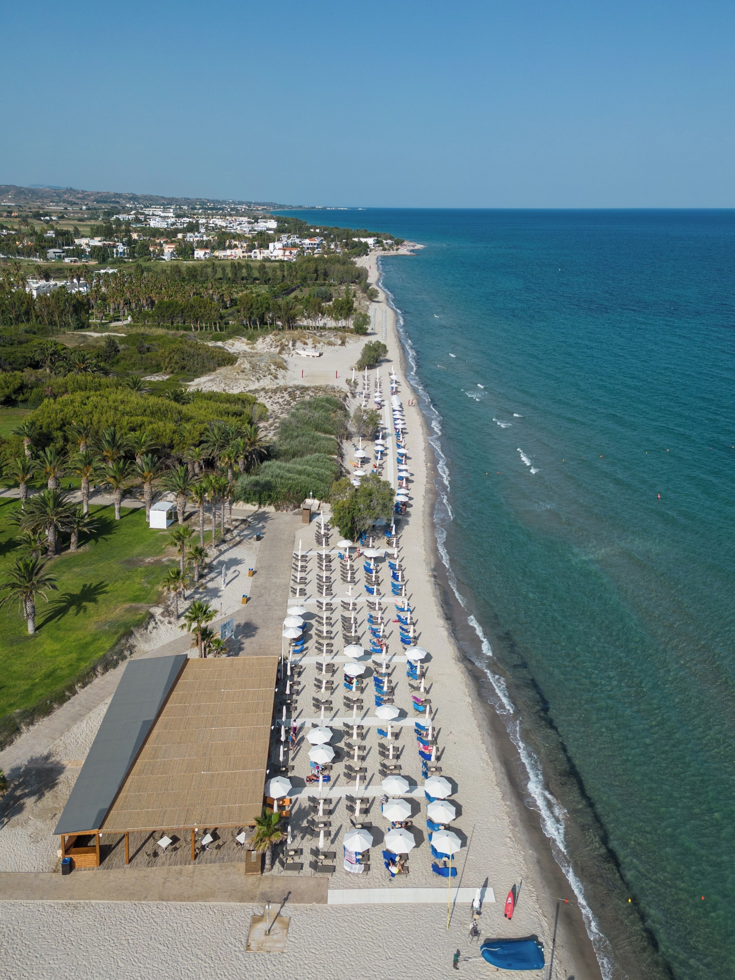 An aerial view of a beach with rows of umbrellas and sun loungers, a clear view of the ocean, and a distant cityscape.