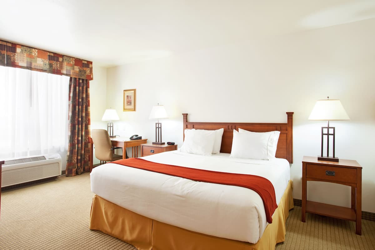 A hotel room with a large bed, two bedside tables with lamps, a desk with a chair, and a window with curtains.