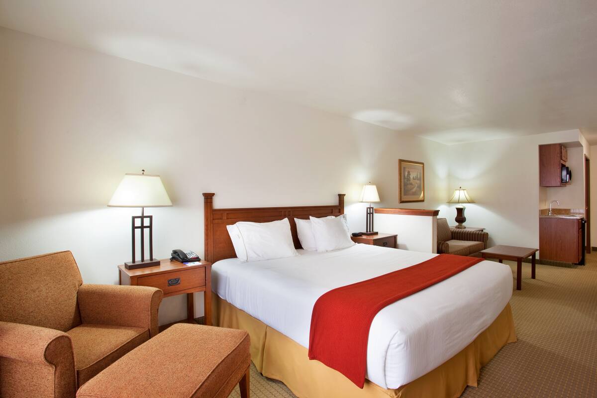 A hotel room with a large bed, two bedside tables, a desk, a chair, a sofa, and a small kitchenette.