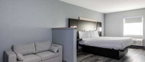 Une chambre d’hôtel moderne avec un canapé gris, un lit avec un cadre noir, un grand miroir et une fenêtre avec des stores.