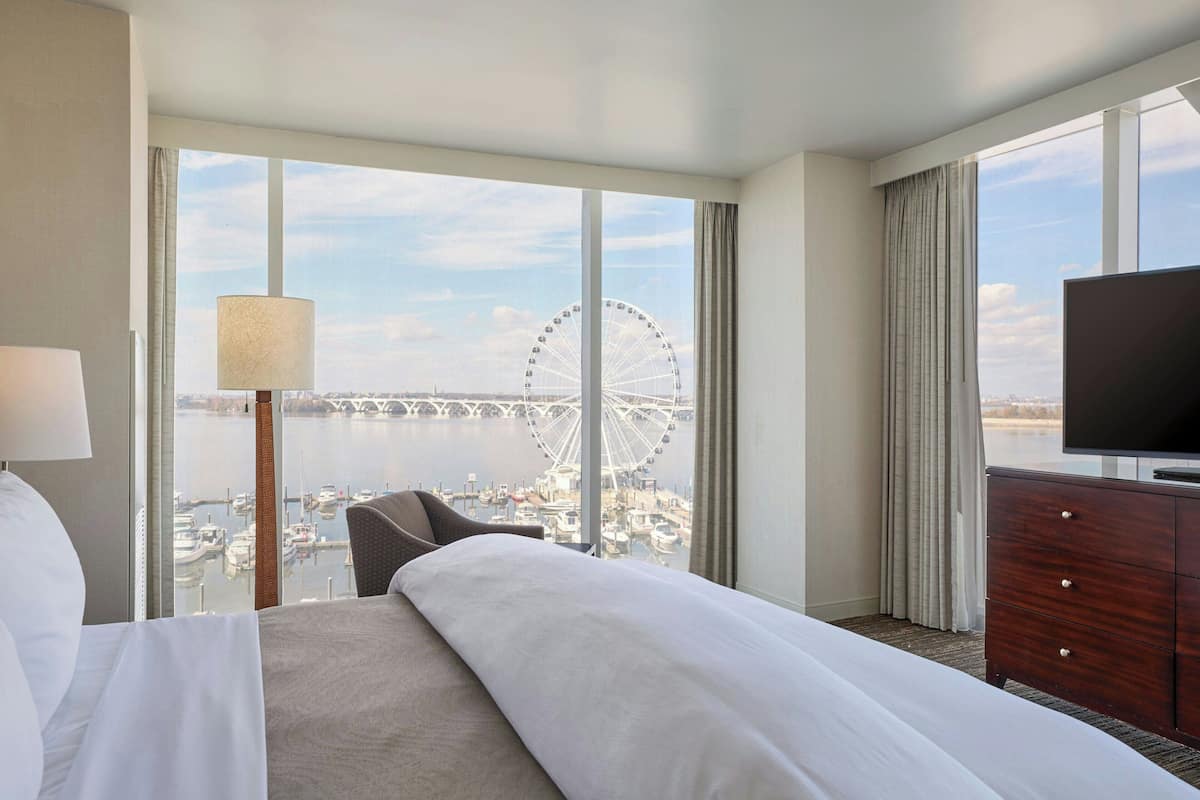 A hotel room with a large bed, a TV, a lamp, and a view of a Ferris wheel and water.