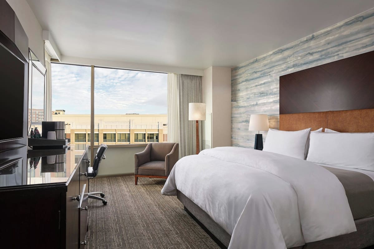 A hotel room with a large bed, a desk with a chair, a television, and a view of the cityscape through large windows.