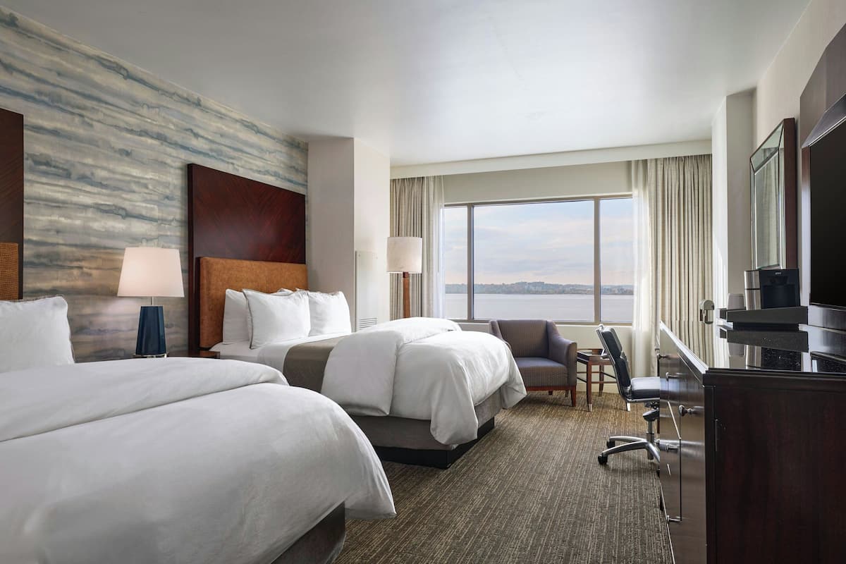 A hotel room with two beds, a desk, a chair, a TV, and a large window with a view.