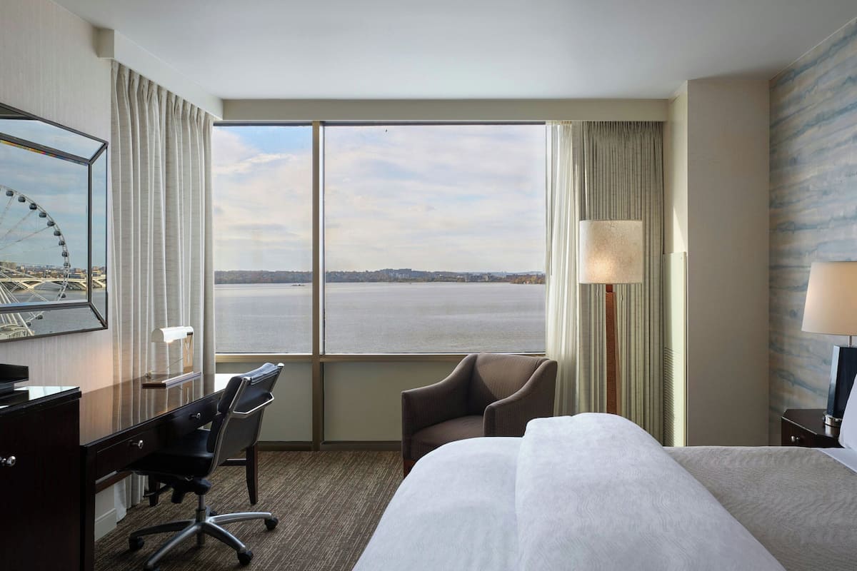 A hotel room with a large bed, a desk with a chair, a lamp, and a view of the water.