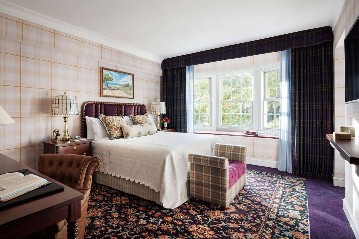 Habitación de hotel con una cama grande, un escritorio con lámpara, una silla, un ventanal con cortinas y una alfombra estampada.