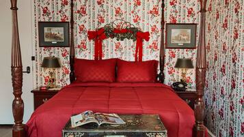 Ein Schlafzimmer mit Himmelbett, roter Bettwäsche, blumigem Wandpapier und je einem gerahmten Bild auf den Nachttischen.