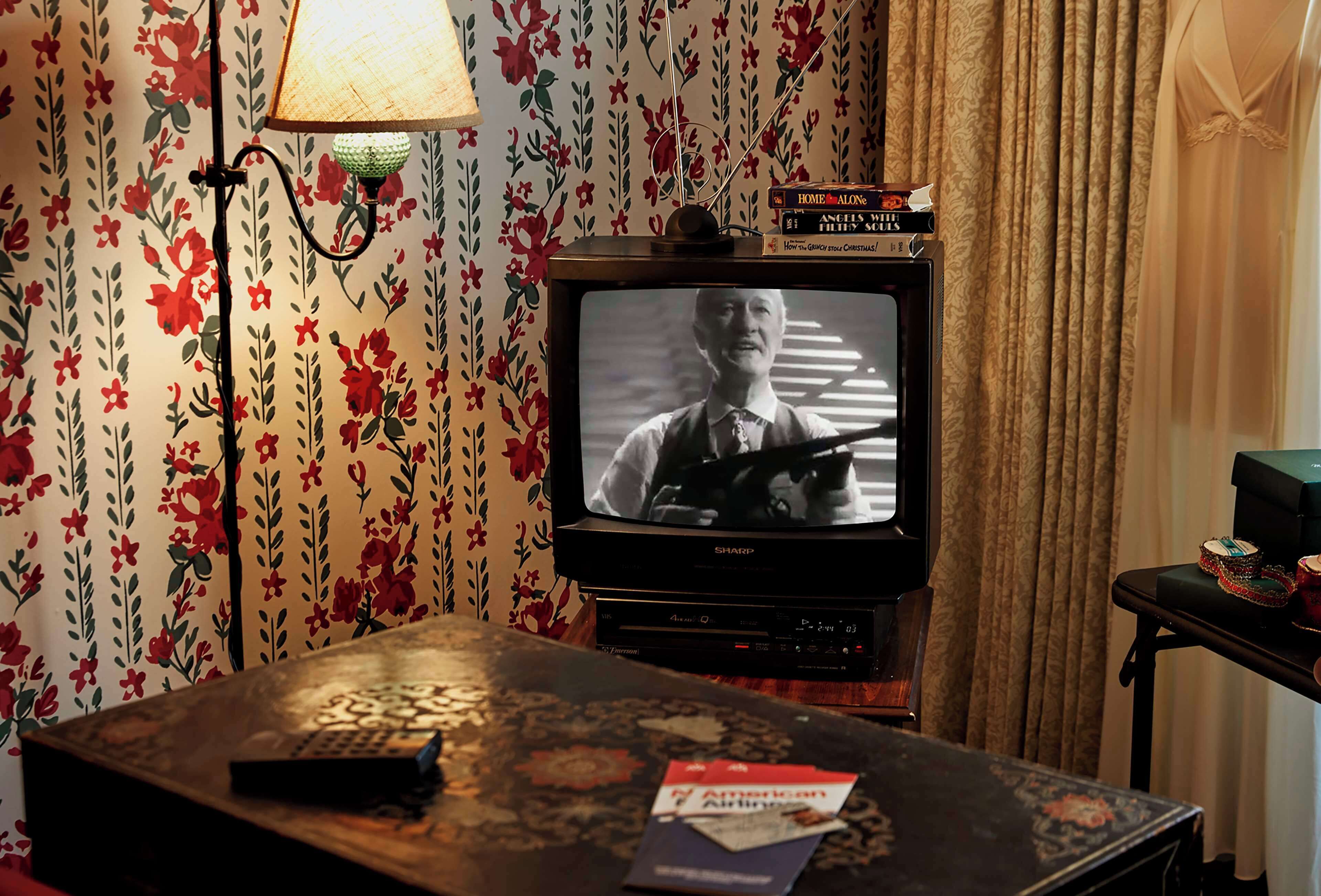 Una habitación con una televisión sobre un soporte, una lámpara y una mesa con un control remoto y una revista.